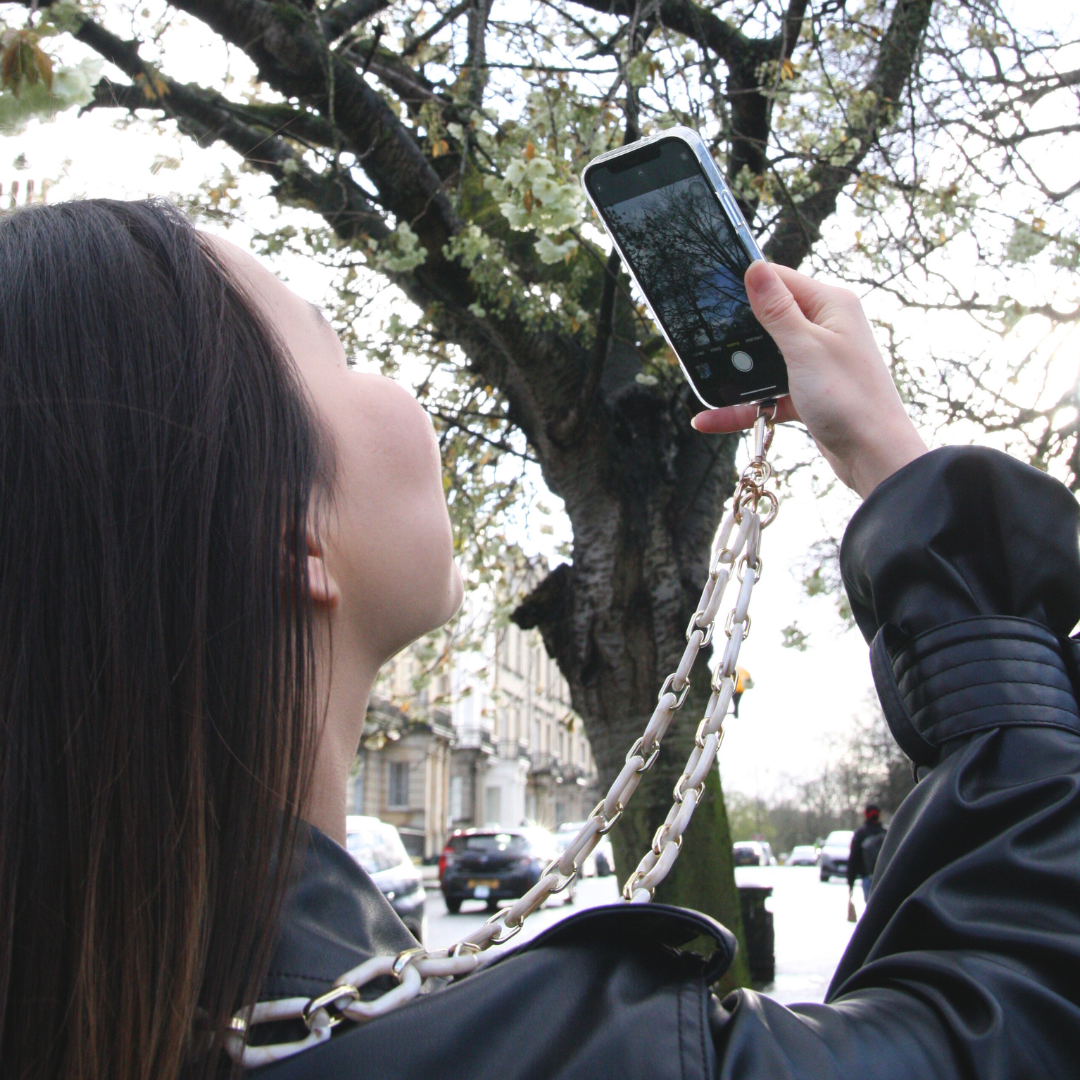 Girl standing outside taking a photo of the tree. Phone is connected with Crossbody phone strap.