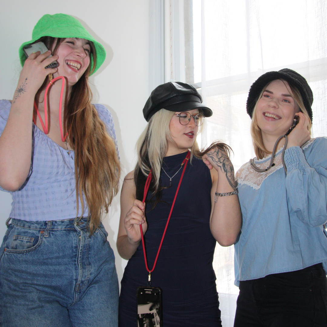 Three girls smiling and posing. Wearing different coloured crossbody phone strap ropes.