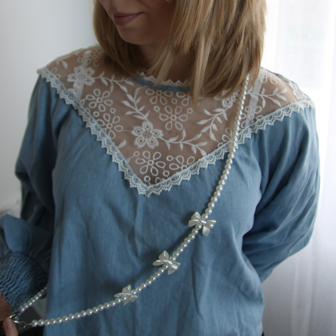 Blond girl wearing blue blouse and wearing a white crossbody phone strap with pearls and bows.