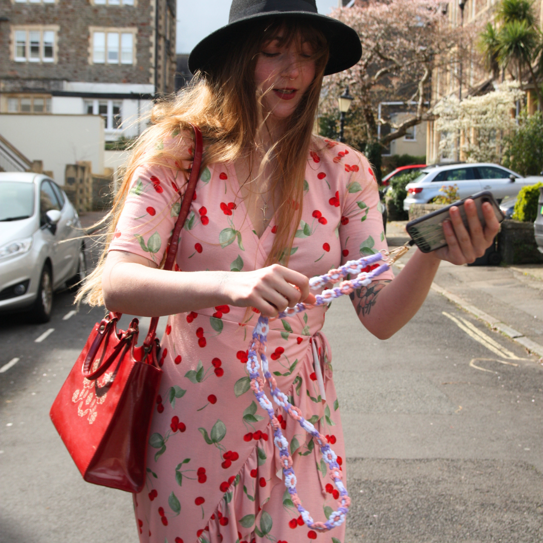 Girl with black hat and brown hair standing in the street, holding her purple, white and pink coloured crossbody phone strap with flowers.